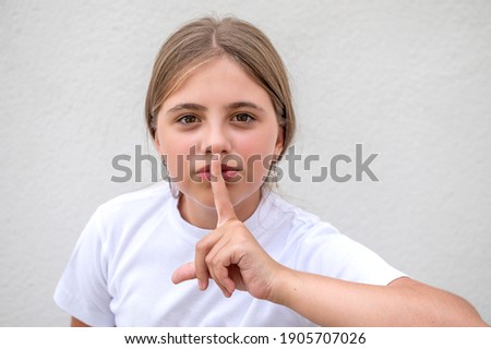 Girl put her finger on mouth keep silent concept. Candid portrait of teenage girl hiking outdoors. Cute girl in a white t-shirt against a white wall.