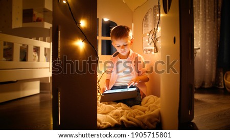 Little smart boy sitting in toy cardboard house and using tablet computer. Concept of child education and studying at night.