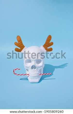 White skull with deer antlers on blue pastel background eats candy cane. Christmas minimal mood concept. New Year’s greeting card.