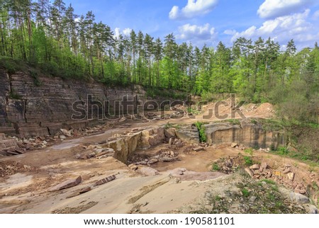 Wide angle view of a quarry with layers of unsurfaced sandstone, and forest and sky in background 