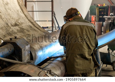a welder at work in an industrial building welds a metal part with his back to us. welding flashes are visible