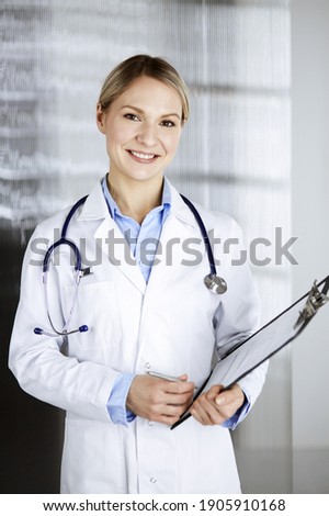 Friendly female doctor standing and holding clipboard in clinic . Portrait of cheerful smiling physician. Perfect medical service in hospital. Medicine and healthcare concept