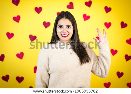 Young caucasian woman over yellow background with red hearts doing hand symbol