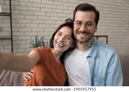 Head shot portrait smiling man wearing glasses and woman hugging, taking selfie, sitting on cozy couch at home, happy couple posing for photo for social network, using gadget, looking at camera