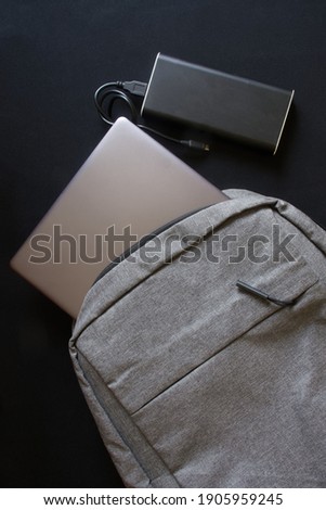 A gray textile city backpack with a silver laptop or ultrabook and an external battery on a black background. Traveling with gadgets. Black gradient