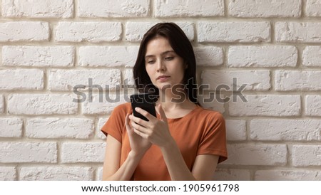 Head shot close up serious young woman using phone, standing on white brick wall background, focused attractive female looking at phone screen, browsing apps, shopping online, reading message