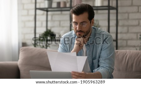 Close up serious man wearing glasses reading letter, payment notification, holding paper, checking financial documents, focused businessman working with correspondence, sitting on couch at home