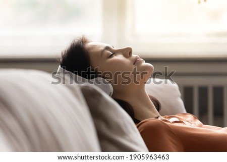Close up peaceful young woman wearing headphones listening to music with closed eyes, leaning back on cozy couch at home, mindful calm sleepy attractive female enjoying favorite song, leisure time Royalty-Free Stock Photo #1905963463