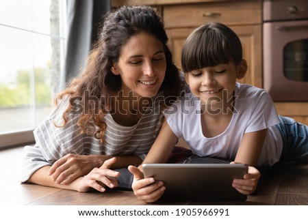 Close up happy mother and adorable little daughter playing tablet, lying on warm wooden floor with underfloor heating, enjoying leisure time with gadget, watching video, chatting or shopping online