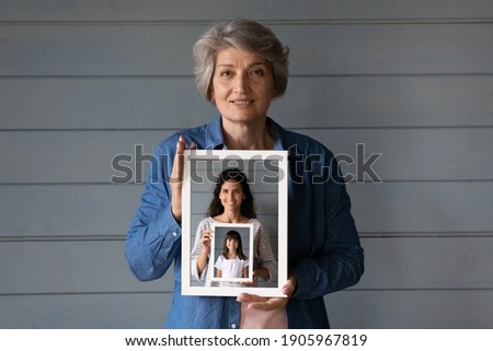 Head shot portrait smiling mature woman holding photo frame of grownup daughter and little granddaughter in row, standing on grey wooden wall background isolated, three generations of women concept