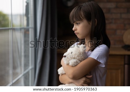 Close up lonely little girl hugging toy, looking out window, standing at home alone, upset unhappy child waiting for parents, thinking about problems, bad relationship in family, psychological trauma Royalty-Free Stock Photo #1905970363