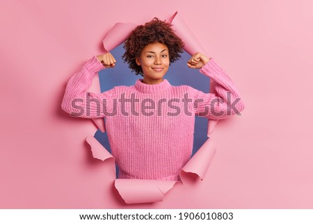Self assured beautiful young Afro American woman raises arms shows biceps being strong and powerful proud of her own achievements wears casual knitted sweater breaks through paper background Royalty-Free Stock Photo #1906010803