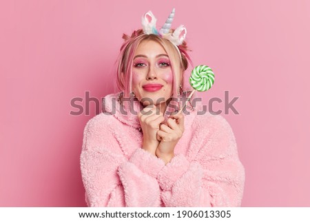 Portrait of pleased young adorable European woman keeps hands near face looks gladfully at camera wears winter coat holds tasty green candy on stick isolated over pink background. Fashion concept