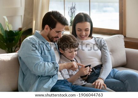 Smiling young Caucasian parents relax on sofa at home with small preschooler son teach using modern cellphone. Happy family with little 6s boy child have fun browsing smartphone. Technology concept.
