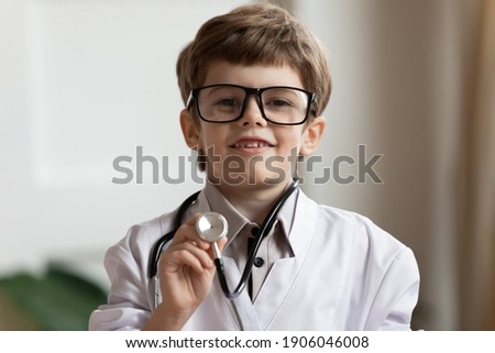 Close up headshot portrait of cute small 7s boy in white medical uniform use stethoscope play hospital. Funny little child have fun act as doctor in clinic, dream of future career. Healthcare concept.