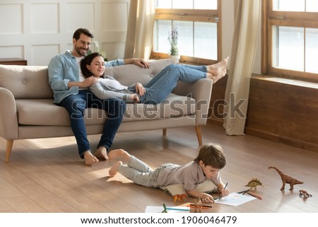 Happy young Caucasian parents relax on cozy couch in living room at home. Small 7s son lying on warm floor drawing painting in album. Overjoyed family with little child rest on weekend in own house.