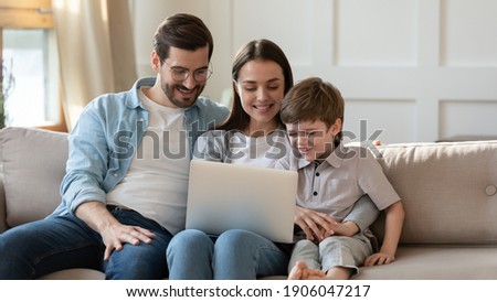 Happy young Caucasian family with little son relax on sofa at home talk on video call on laptop. Smiling parents with small 7s boy child look at computer screen watch funny cartoon online on gadget.