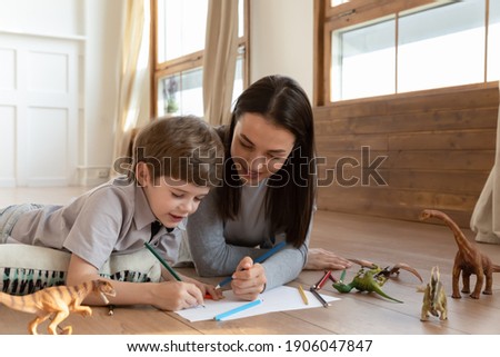 Happy young Caucasian mom and small 6s son lying on floor at home have fun drawing together in album. Smiling loving mother or nanny involved in creative activity painting. Hobby concept.