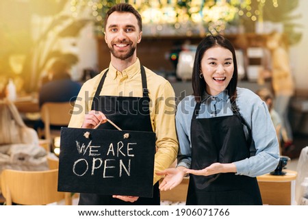 Portrait of a two successful friendly waiters. Satisfied young man and pretty asian young woman in black aprons stand inside a restaurant, cafe or bar, showing signboard OPEN, with pleasured smiling
