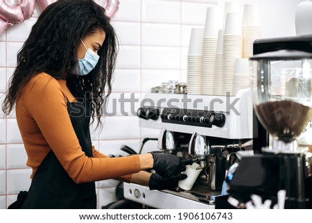 Young African American female barista wearing a medical mask and black apron makes coffee for a coffee shop visitor at their. Owner of small business. Cafe, service industry concept