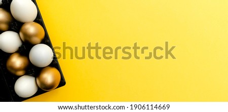 Gold and white easter eggs in a black egg holder. Bright yellow background banner. Top horizontal view.