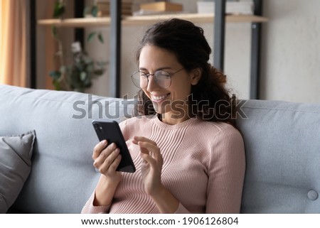 Happy millennial woman relaxing on comfortable sofa, enjoying web surfing information in internet or using mobile software application, communicating distantly with friends in social networks at home.