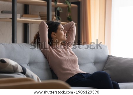 Relaxed young smiling pretty woman sitting on cozy couch, looking in distance, dreaming or visualizing future, recollecting memories, enjoying stress free peaceful weekend holiday time at home. Royalty-Free Stock Photo #1906129390