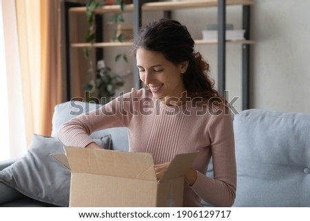 Smiling attractive woman opening big carton box, feeling curious of unpacking internet store order at home. Happy millennial female client satisfied with fast delivery service, online shopping concept Royalty-Free Stock Photo #1906129717
