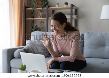 Happy young woman in eyeglasses looking at computer screen, starting online video call conversation with friends, family or colleagues, sitting on sofa at home, modern tech and distant communication. Royalty-Free Stock Photo #1906130293
