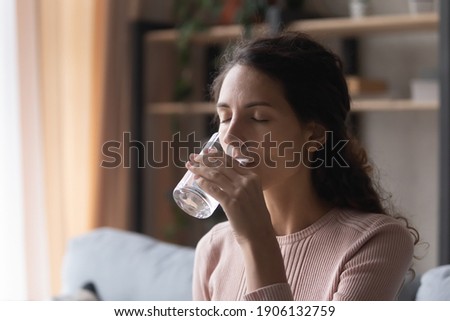 Head shot happy young caucasian woman drinking glass of fresh pure mineral water, sitting on comfortable sofa at home. Peaceful millennial lady enjoying healthcare morning daily habit indoors. Royalty-Free Stock Photo #1906132759