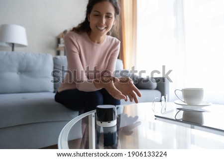 Happy young woman using wireless voice assistant small modern technology gadget, listening music. Smiling female client satisfied with stylish compact portable speaker at home, searching in internet.