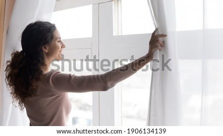 Smiling young dreamy woman opening curtains, looking outside window in morning. Sincere millennial caucasian lady breathing fresh air, starting new day or enjoying peaceful weekend alone at home. Royalty-Free Stock Photo #1906134139