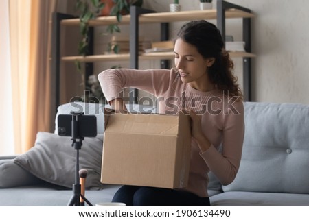 Smiling pretty young female influencer unpacking big cardboard box, sitting in front of mobile phone on stabilizer, sharing internet store shopping experience with followers online or recording video. Royalty-Free Stock Photo #1906134490