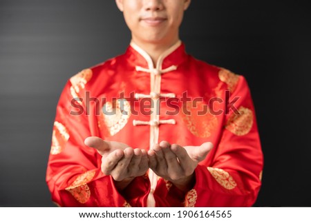 Showing holding empty palm hand of happy positive handsome young Asian man in red Chinese clothes, Qipao or Cheongsam is showing product with copy space to text with fade shade black background.