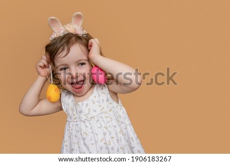 Close-up Emotional Positive portrait of little girl with bunny ears on her head, plays an orange pink egg as earrings on her ears. Isolated light orange background copy space, postcards. Festive child