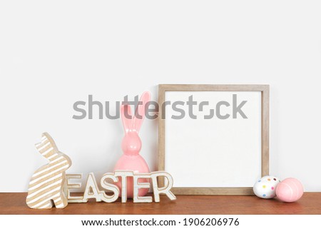 Mock up wood frame with Easter decorations on a wood shelf. Rustic wood decor, eggs, modern glass bunny. Square frame against a white wall. Copy space.