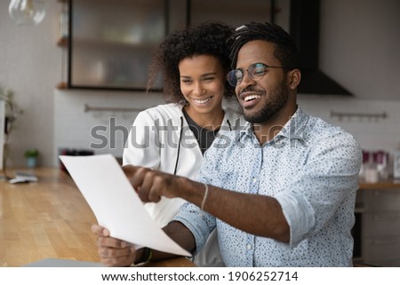 Happy young black married couple reading paper letter about financial reward at kitchen. Smiling satisfied afro american husband wife discuss good conditions of loan mortgage proposal received by mail Royalty-Free Stock Photo #1906252714