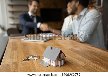 Focus on close up keys bunch and cottage house toy model on wooden table. Smiling young man broker realtor real estate agent shake hands of happy black couple clients homeowners on blurred background Royalty-Free Stock Photo #1906252759