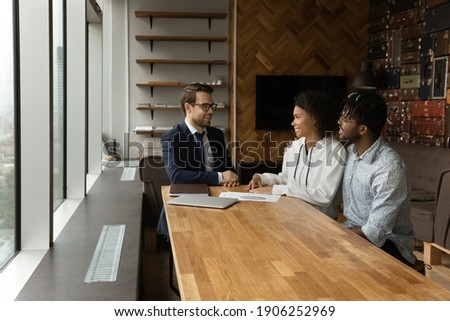 Happy african american couple sit by desk at travel agent office listen to pleasant male consultant choose trip. Confident young businessman talk with black husband wife customers offer house to buy Royalty-Free Stock Photo #1906252969