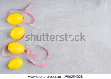 Top view of five painted in vibrant yellow color eggs prepared for easter party laying on gray concrete background in a line at the left side with pink ribbon. Image with copy space, horizontal