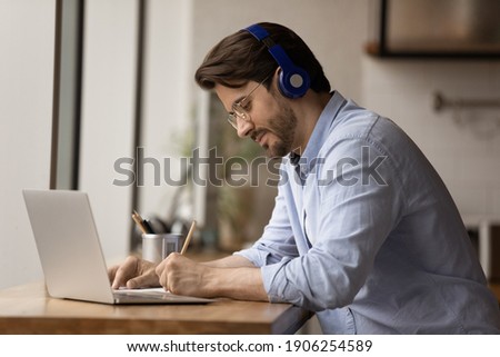 Diligent millennial man remote student sit by pc in earphones listen to teacher tutor questions write answers on paper by hand. Busy young male interpreter translate foreign audio material make notes
