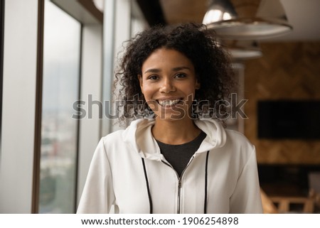 Headshot portrait of beautiful smiling millennial afro american female in casual clothing posing at modern studio apartment. Happy young black lady looking at camera. Profile picture at social network