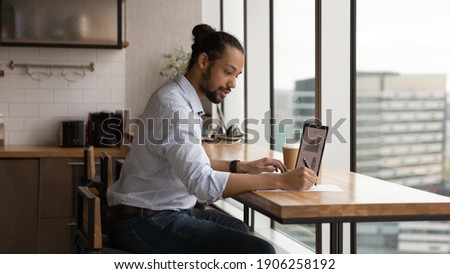 Focused young biracial man sales manager prepare summary report at home office check stats on pc screen make records on paper. Black male financial expert study stock prices rate growth graphs online Royalty-Free Stock Photo #1906258192