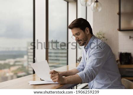 Successful businessman read paper document at work desk in studio apartment near panoramic window. Thoughtful confident young man freelancer engaged in paperwork at home office study official letter Royalty-Free Stock Photo #1906259239