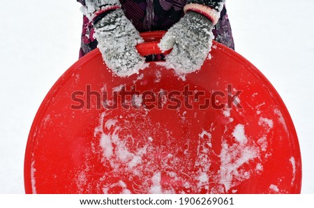 The hands of a child in snow-covered mittens hold a red ice box all in the snow. Winter fun-skiing down the mountain.