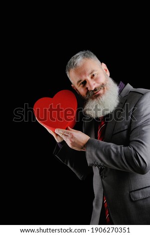 Mature bearded man wear gray suit holds red heart shaped gift box, isolated black background