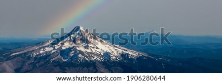 A summer panoramic aerial shot of Mt Hood with rainbow with much of its snow melted and showing layered hills and Mt Jefferson in background