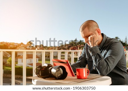 Young photographer sitting with a coffee, at the table on the terrace of his house, looking at the tablet screen and learning photography. studying Online education. working at home. worried and tired