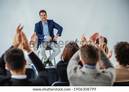 Smiling motivational speaker sitting in front of his audience who is clapping. Royalty-Free Stock Photo #1906308799