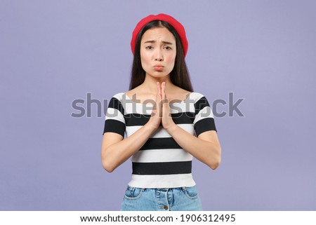 Pleading begging young brunette asian woman 20s wearing striped t-shirt red beret standing holding hands folded in prayer looking camera isolated on pastel violet colour background studio portrait
